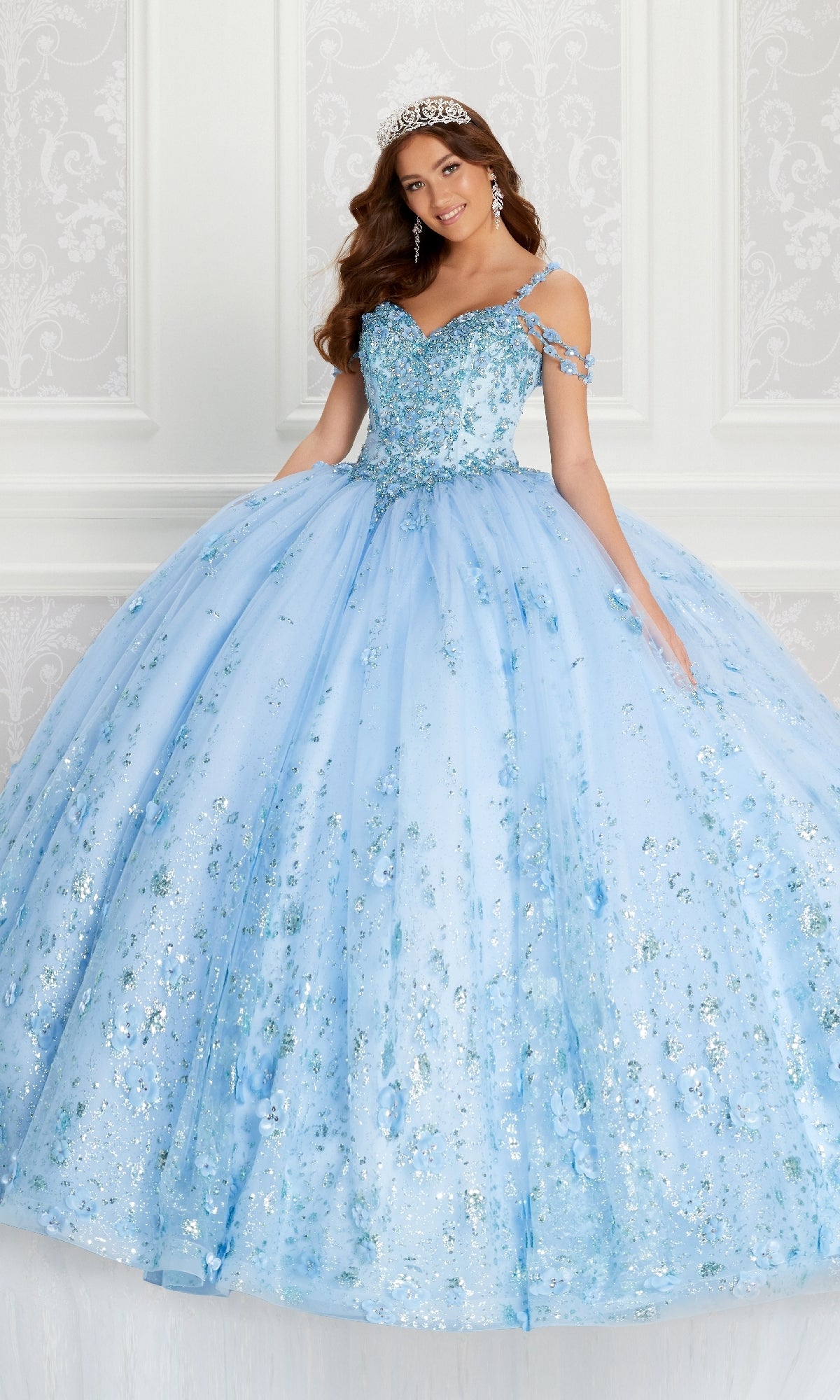 A woman wearing the Quinceanera Dress PR22145 By Princesa—a sparkling light blue off-the-shoulder gown with rhinestone embellishments and a tiara—stands in front of an ornate white wall.
